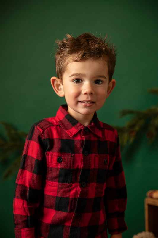 Young boy in red buffalo check shirt in Christmas clothes sitting in studio set with green background. Gin Quist Photography