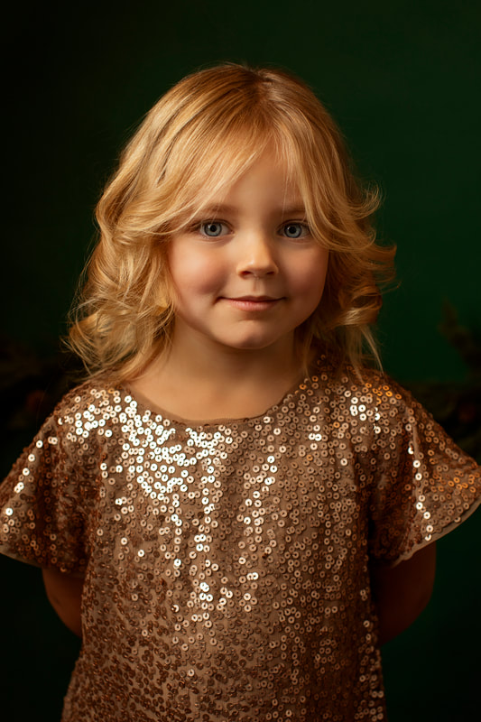 Photo of small girl with small smile in sparkling dress with dark green background. Gin Quist Photography